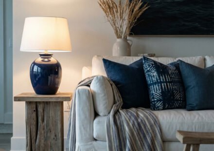White slipcovered sofa with navy pillows, striped throw, blue ceramic lamp, ocean art, and jute rug creating a refined coastal summer aesthetic in a serene living room.