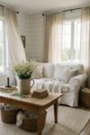 Farmhouse Summer Decor That Feels Light And Lived In White slipcovered sofa, linen curtains, rustic wood coffee table, daisies in a crock, books, and woven baskets create a soft farmhouse summer decor living room.