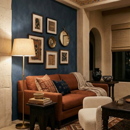 Rust sofa against a deep blue accent wall with framed art, brass floor lamp, patterned rug, and carved wood tables in a warm Nubian Style living room.