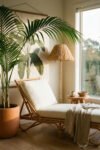 Sunlit corner with rattan chaise lounge, large potted palm, woven floor lamp, small wood stool, and neutral textiles for relaxed summer decor.