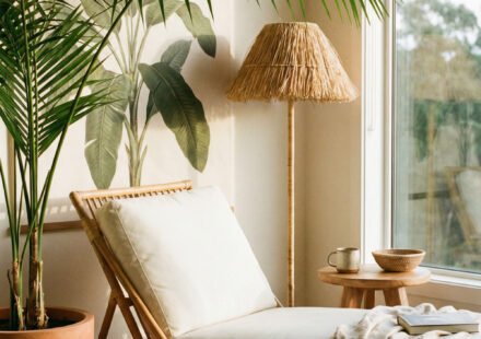 Sunlit corner with rattan chaise lounge, large potted palm, woven floor lamp, small wood stool, and neutral textiles for relaxed summer decor.