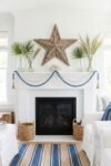 Coastal fireplace mantel with a driftwood star, blue and white bead garland, palm leaves, coral accent, and striped rug for breezy summer mantle décor inspiration.