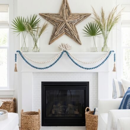 Coastal fireplace mantel with a driftwood star, blue and white bead garland, palm leaves, coral accent, and striped rug for breezy summer mantle décor inspiration.