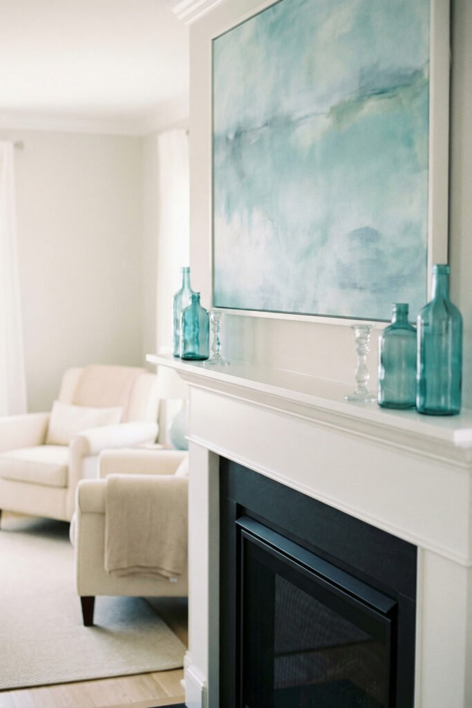 White mantel with blue abstract art, aqua glass bottles, and clear candlesticks for modern coastal summer mantle décor.
