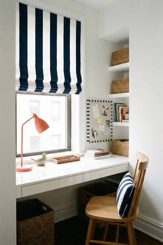 Built-in white desk with blue striped roman shade, coral lamp, shelves, and wicker baskets creating crisp Summer Office Decor in a tiny office.
