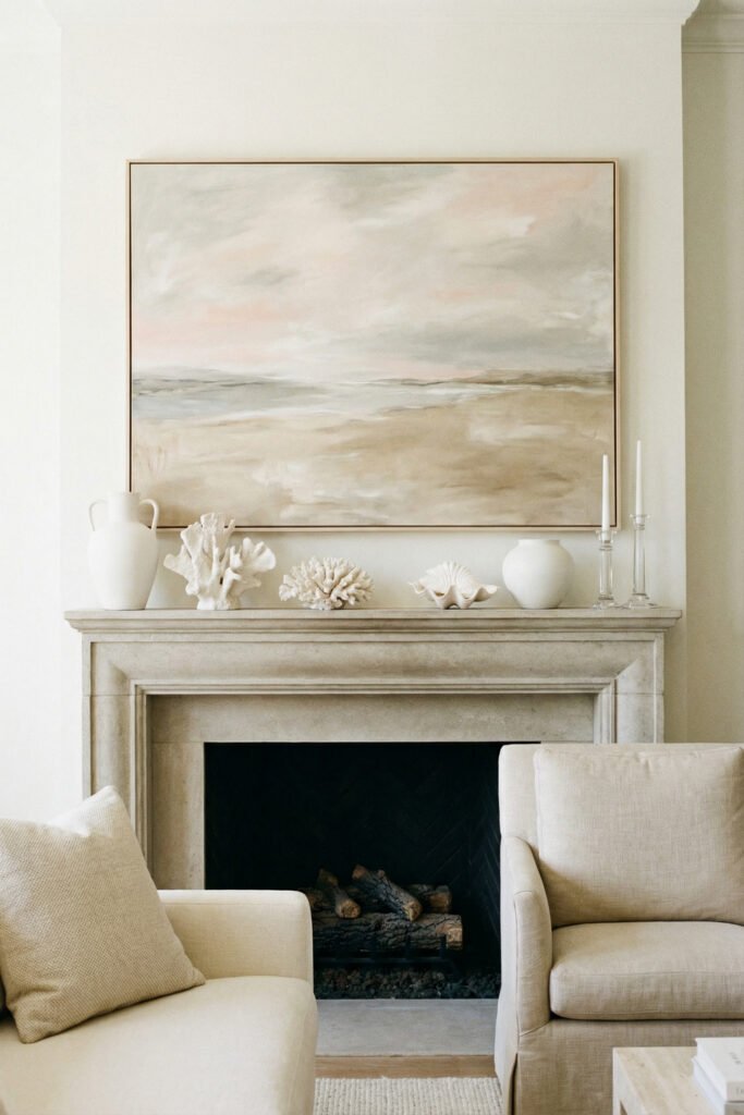 Classic stone mantel with muted coastal artwork, white ceramics, coral accents, and glass candlesticks for elegant neutral summer mantle décor.
