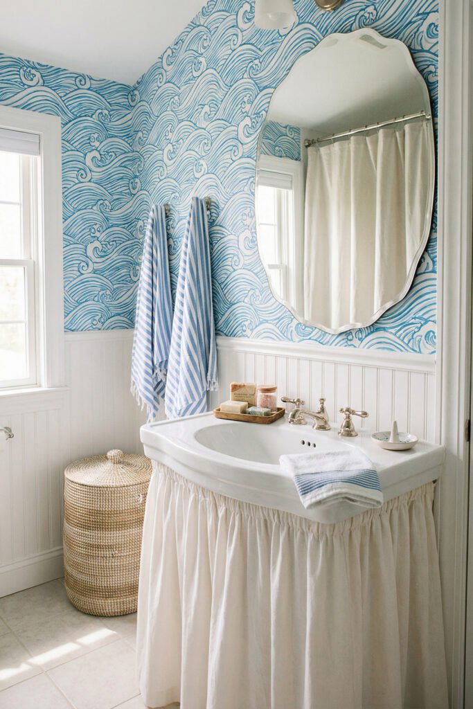 Beach Cottage Decor That Feels Fresh for Summer 16 Bathroom with blue wave wallpaper, white beadboard, skirted sink, striped towels, and wicker hamper styled in fresh beach cottage decor.
