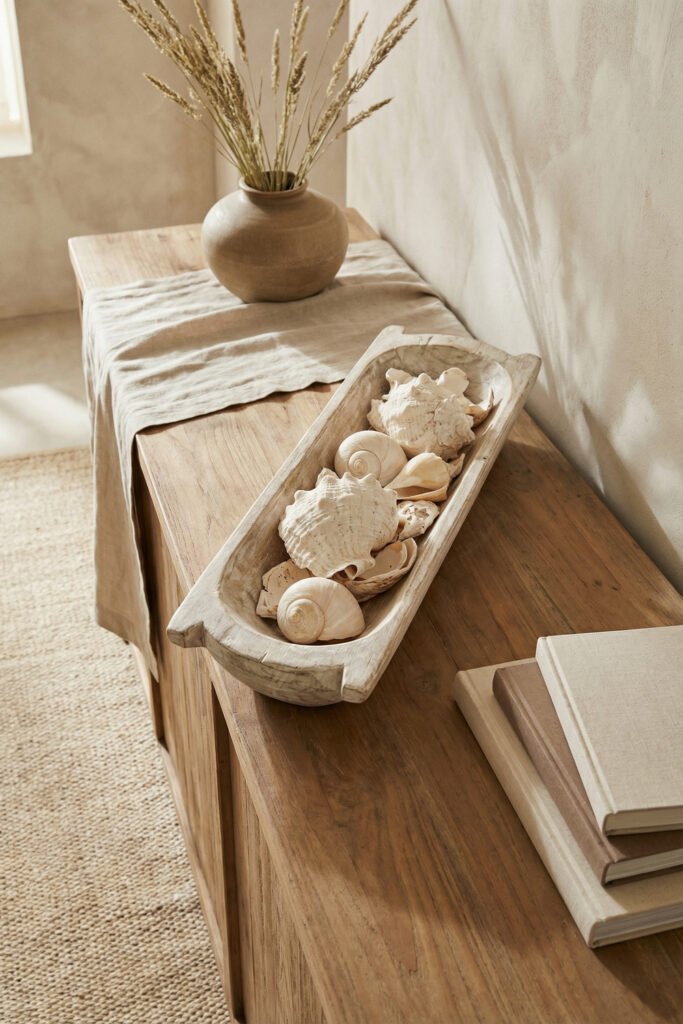 Long rustic wooden bowl filled with large white shells and spiral shells on a warm wood console with linen runner and vase for a natural seashell display.
