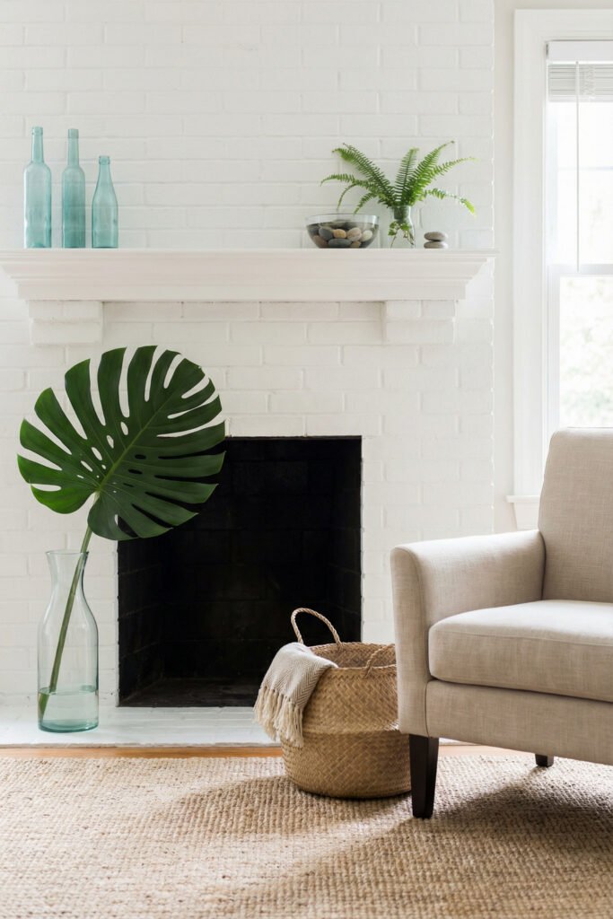 White brick fireplace with aqua bottles, a fern, stacked stones, and a giant monstera leaf in glass for minimal tropical summer mantle décor.
