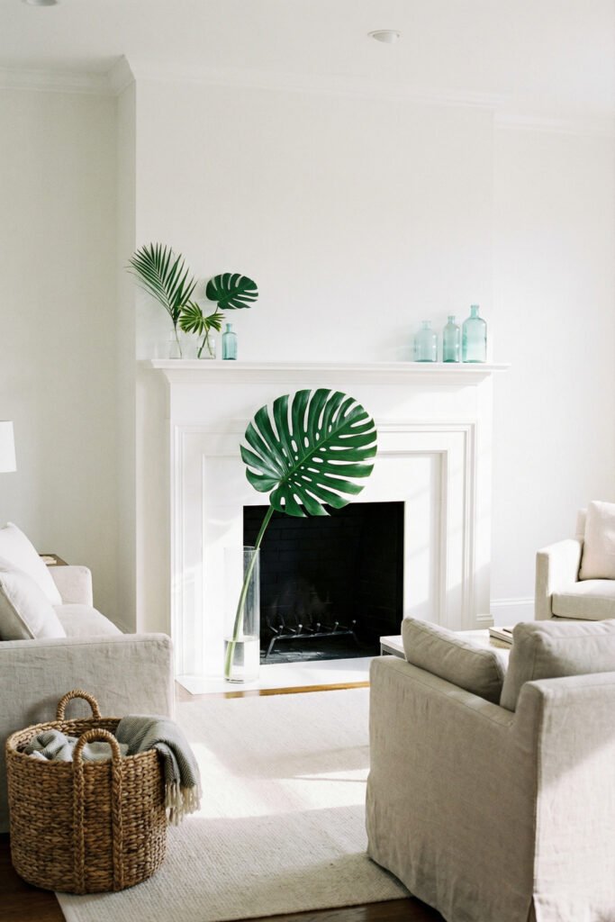 Bright white fireplace styled with monstera and palm leaves plus pale aqua bottles for clean, modern summer mantle décor with tropical flair.
