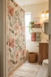 Small bathroom with a watercolor floral shower curtain, warm wood vanity, round mirror, coral and sage towels, wicker basket, and cream rug.