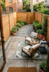 Narrow fenced backyard with cushioned lounge chairs, a patterned rug, round tables, potted plants, gravel borders, and a stepping-stone path.