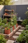 Compact garden with fabric grow bags, a wire trellis, brick-edged planting areas, a stepping-stone path, and a wooden potting bench.