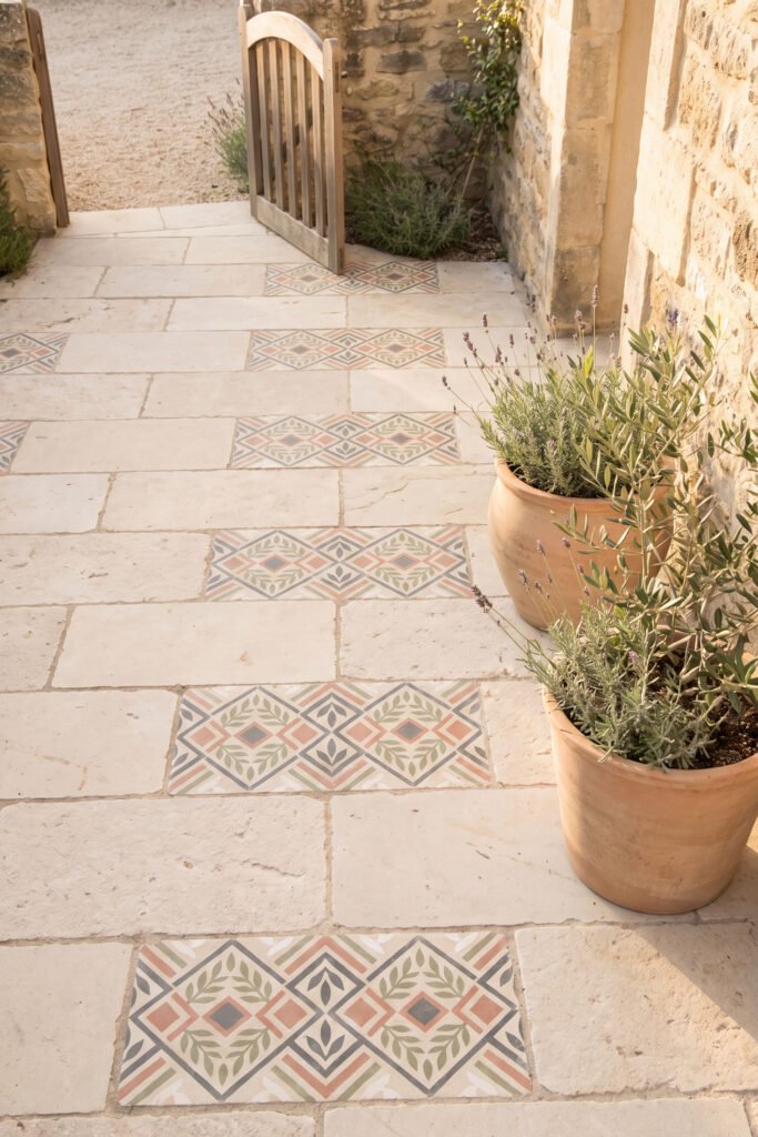 Pale stone courtyard path with evenly spaced stenciled geometric panels, terracotta planters, and an open wooden gate.