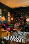 Black salon with a chinoiserie cabinet, burgundy sofa, mustard chair, zebra rug, candles, flowers, and green accents.