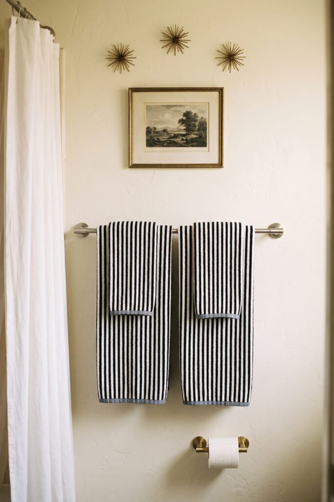 Two black-and-white striped towels hang beneath a framed landscape and three brass starburst accents on a cream bathroom wall.