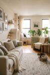 Cream living room with a mustard chair, pale wood cat steps, window perch, woven hideaway, sage screen, leafy plants, and two cats.