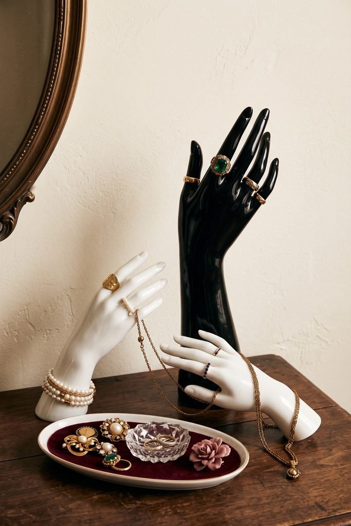 One black and two white sculptural hands display rings, bracelets, and necklaces above a burgundy-lined tray of brooches.
