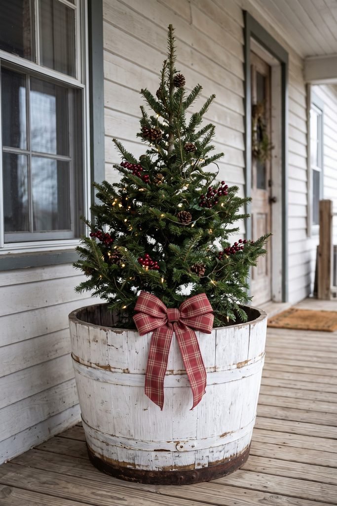 Evergreen tree decorated with pinecones, red berries, warm lights, and a large plaid bow in a whitewashed barrel on a porch.