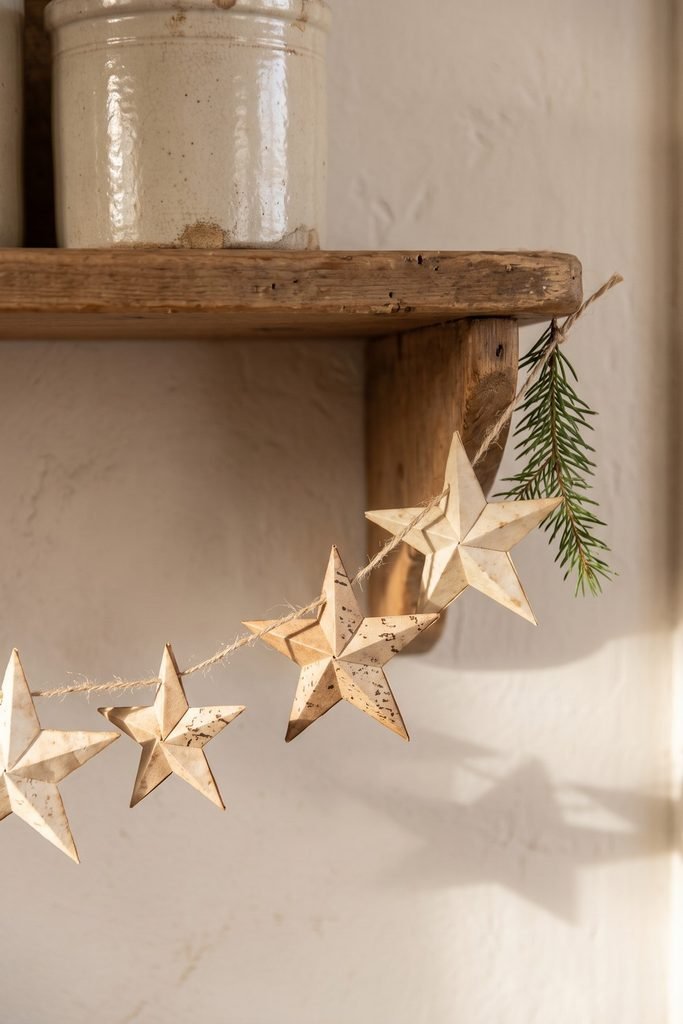 Folded paper stars strung beneath a wooden shelf holding a cream crock, with an evergreen sprig at one end.
