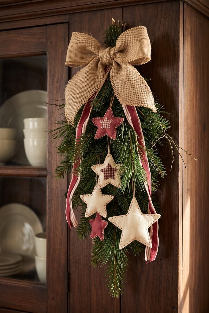 Evergreen swag with stuffed cream and red stars, checked ribbon, and a large burlap bow hanging on a wooden cupboard.